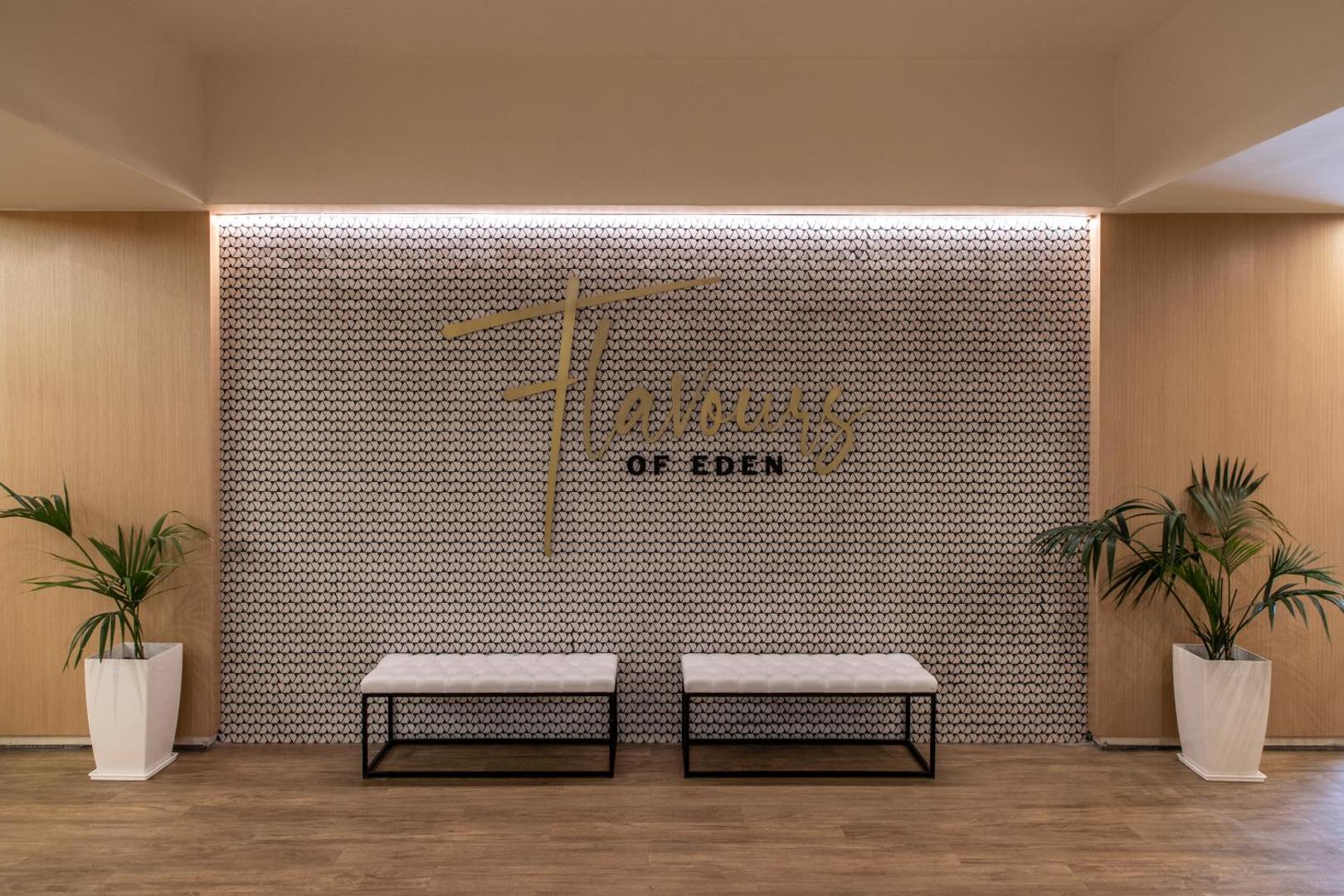 Eden Roc Resort restorāna Flavours of Eden ieeja ar modernu interjeru, dekoratīvu sienu, baltiem soliņiem un telpaugiem podos
