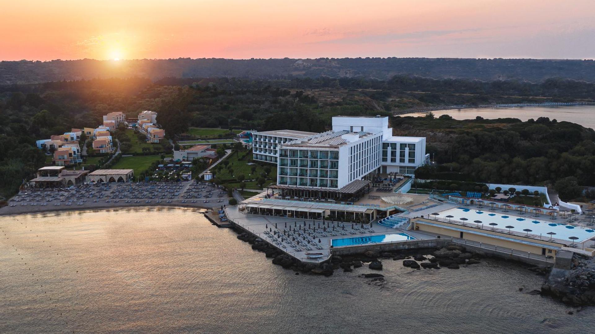 Eden Roc Resort panorāmas skats saulrietā Rodas salā — viesnīcas ēkas, baseini un privātā pludmale pie Egejas jūras līča