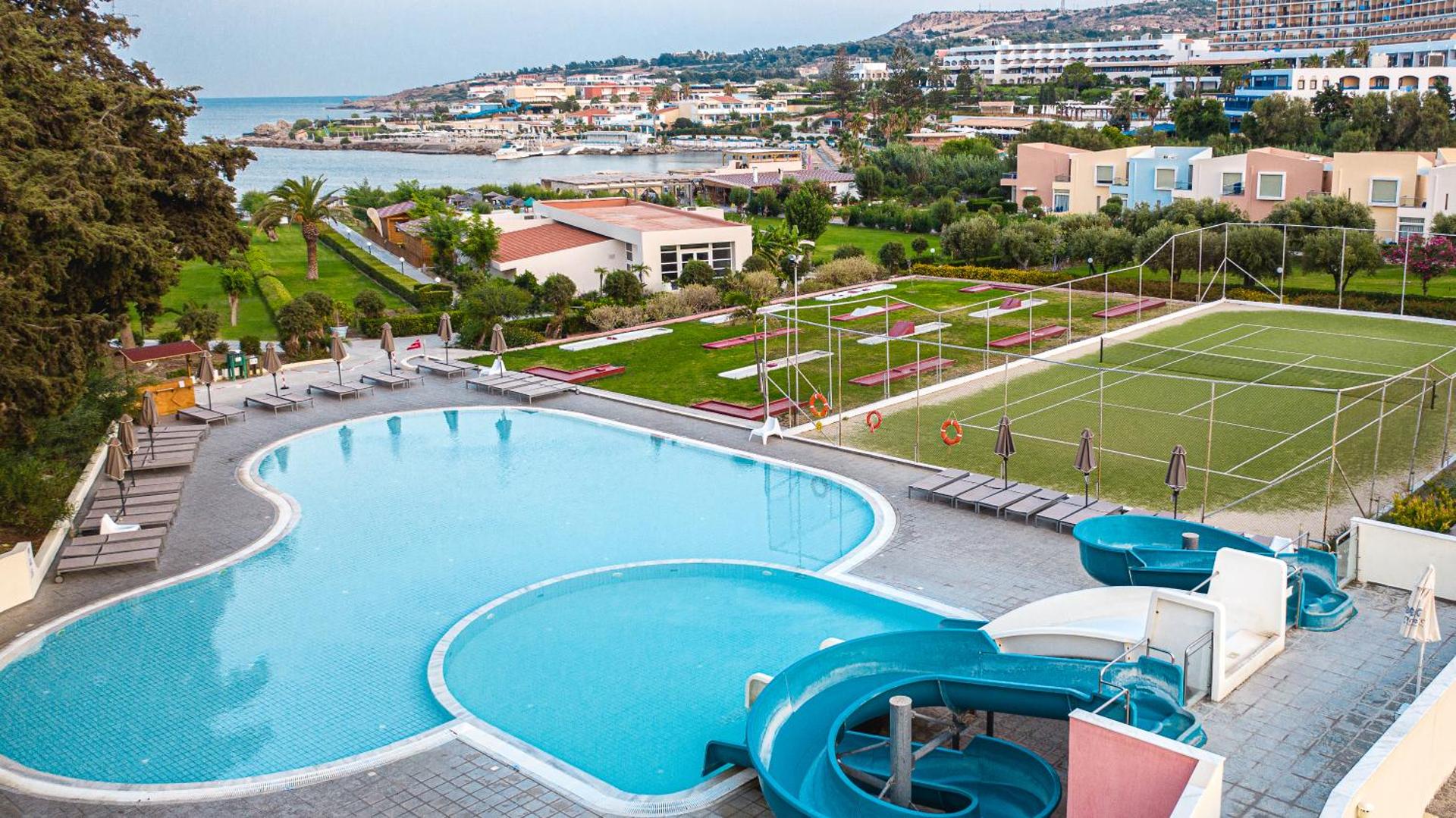 Eden Roc Resort teritorijas skats no augšas ar baseinu, ūdens slidkalniņiem un tenisa kortu piekrastē Rodas salā