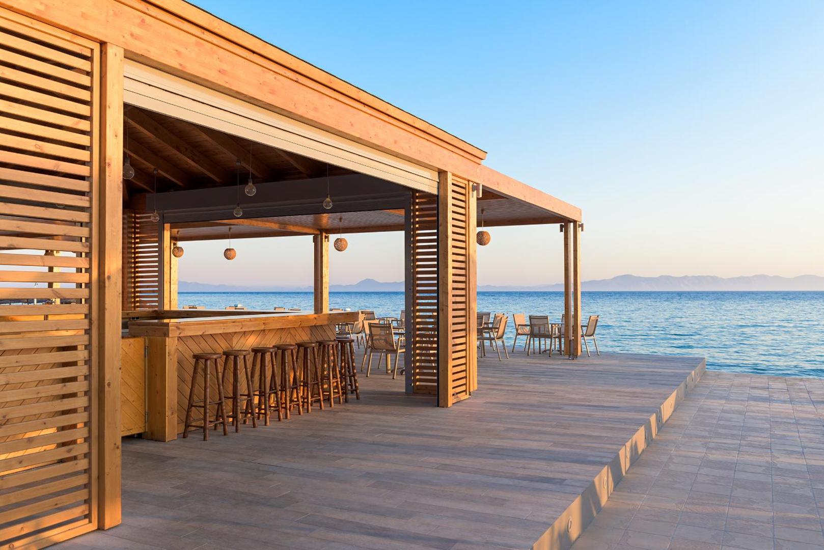 Avra Beach Resort koka terases bārs pie pašas jūras krasta ar bāra krēsliem un galdiņiem saulrieta gaismā Rodā