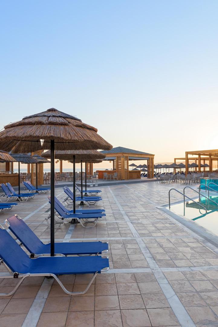 Avra Beach Resort saulessargi un sauļošanās krēsli pie baseina saulrieta laikā, fonā redzams pludmales bārs un jūra