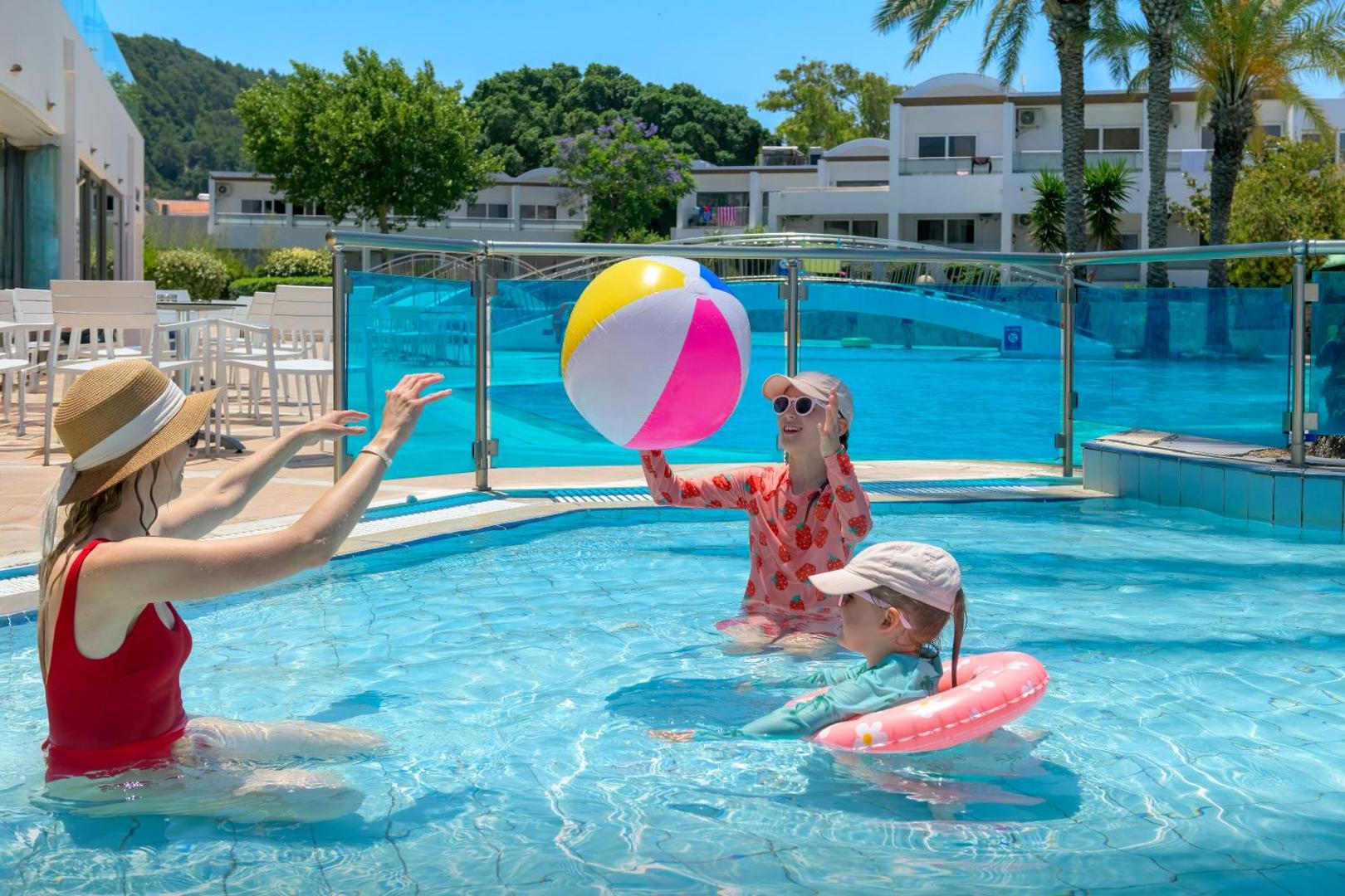 Ģimene ar bērniem spēlējas ar piepūšamo bumbu Avra Beach Resort seklajā baseinā saulainā dienā Rodas salā