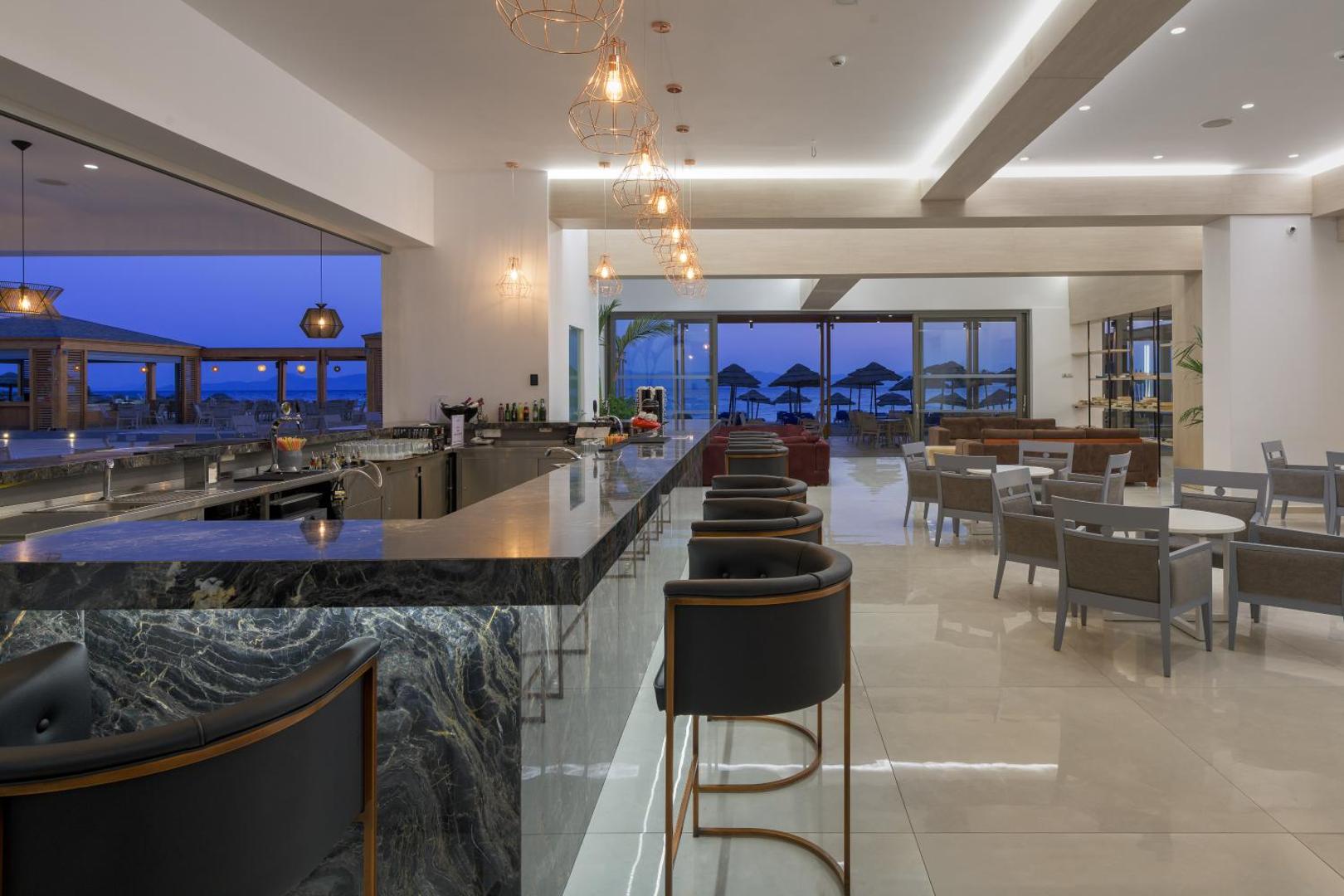 Viesnīcas Avra Beach Resort iekštelpu bārs ar marmora apdari un elegantiem krēsliem, fonā redzama pludmales zona krēslas stundā