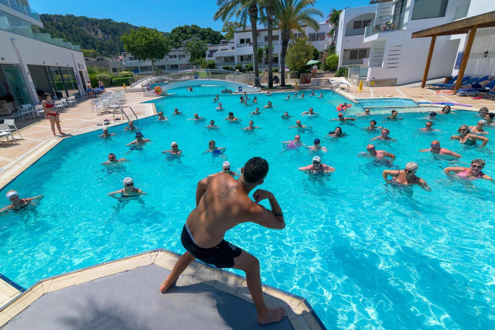 Ūdens aerobikas nodarbība Avra Beach Resort lielajā baseinā trenera vadībā, fonā viesnīcas korpusi un palmas