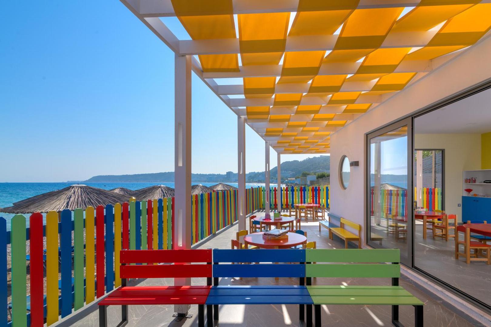Avra Beach Resort bērnu rotaļu laukums uz terases ar krāsainu sētu un nojumi — tiešs skats uz pludmali un jūru