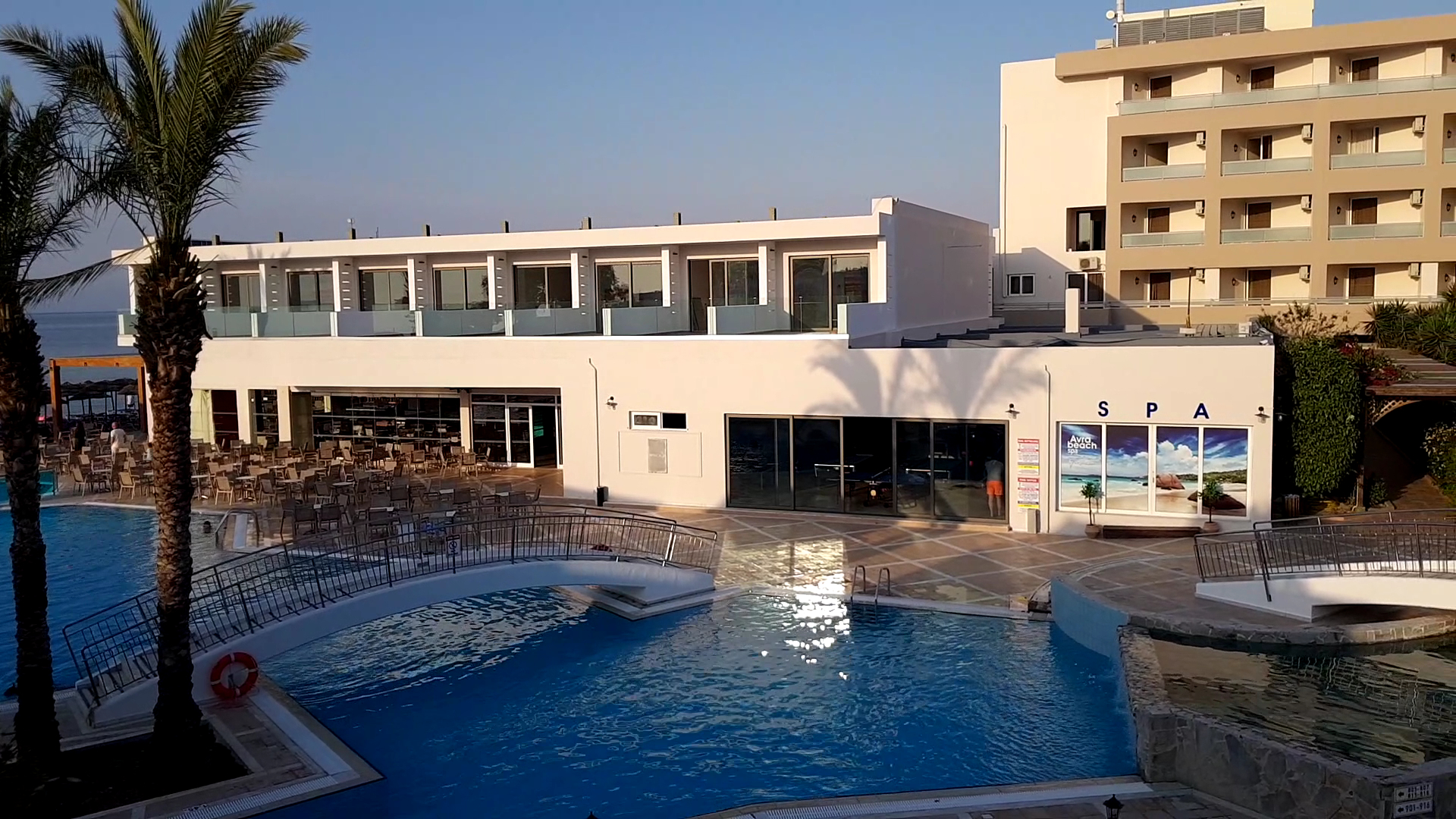 Avra Beach Resort spa centra ieeja un āra baseins ar dekoratīvu tiltiņu — skats uz viesnīcas korpusu un palmu