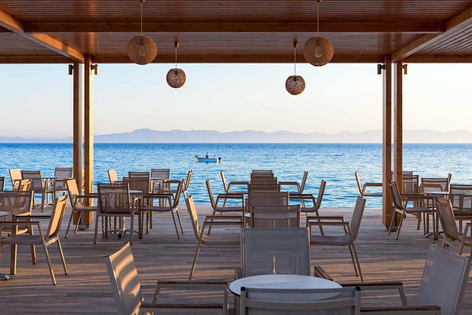 Avra Beach Resort Hotel & Bungalows restorāna terase ar skatu uz jūru Rodas salā — koka nojume, stilīgi gaismekļi un galdiņi piekrastē