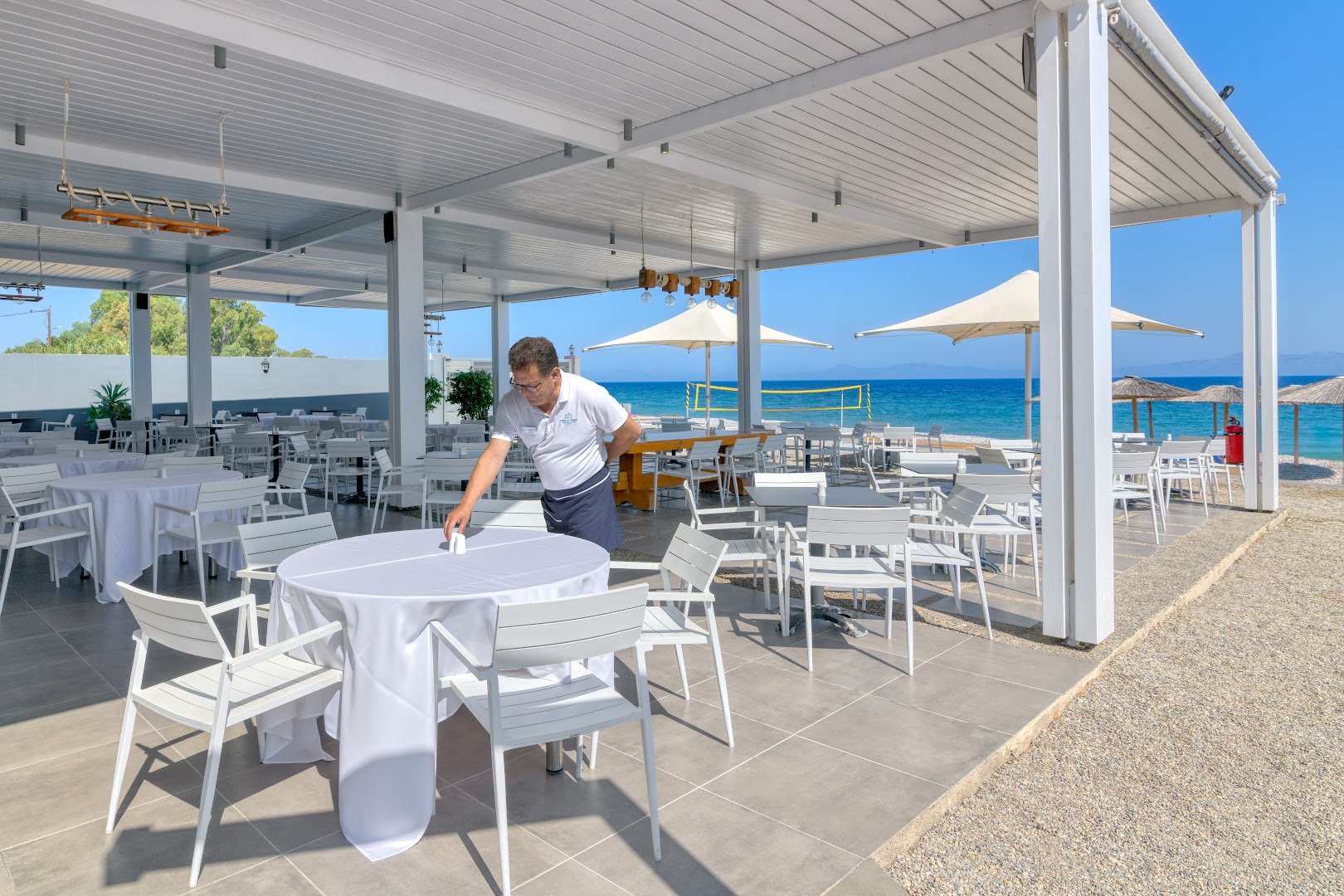 Avra Beach Resort āra restorāns pludmales zonā — balti galdi un krēsli zem nojumes, skats uz oļu pludmali un zilo jūru