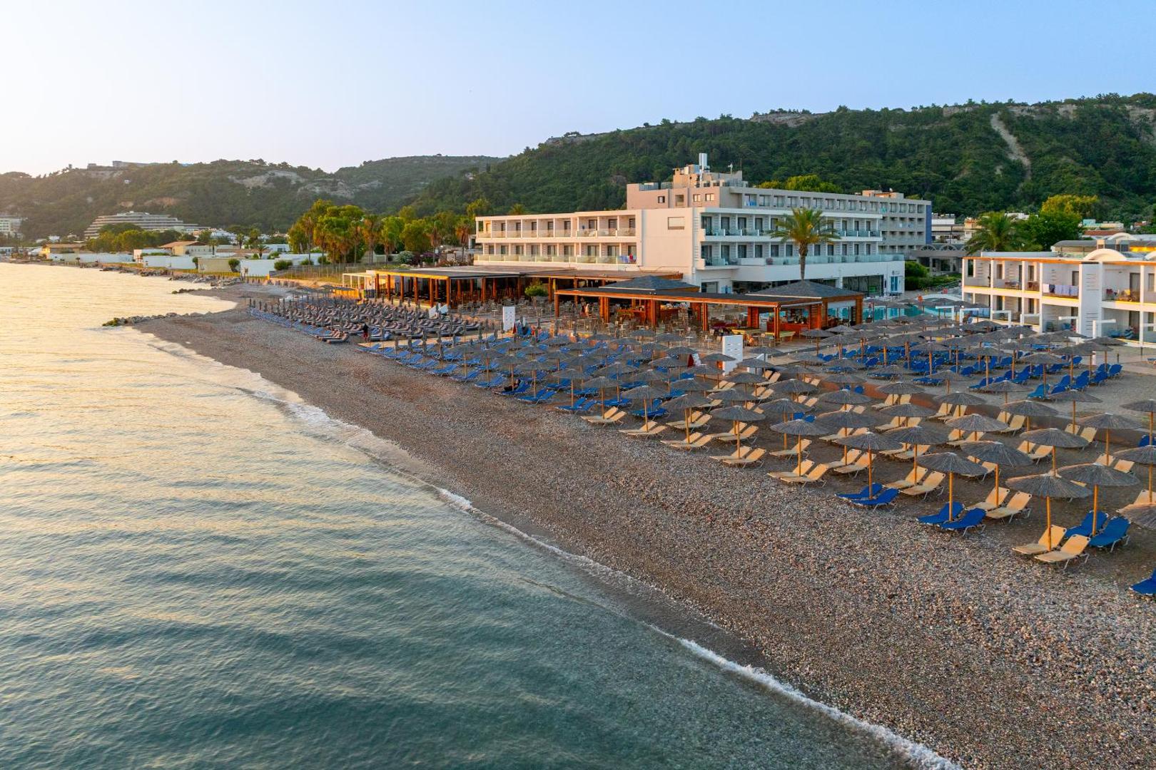 Avra Beach Resort pludmale saulrietā ar daudziem saulessargiem un zviļņiem Rodas salā — skats uz viesnīcas ēku no jūras puses