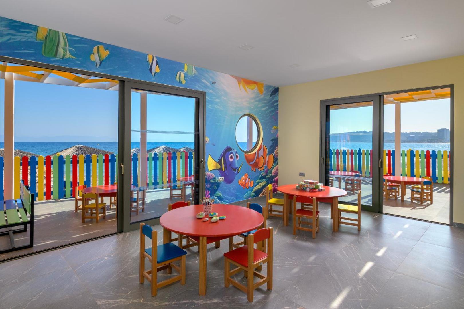Avra Beach Resort bērnu kluba iekštelpas ar krāsainiem galdiem un zīmējumiem uz sienām — skats caur logu uz jūru