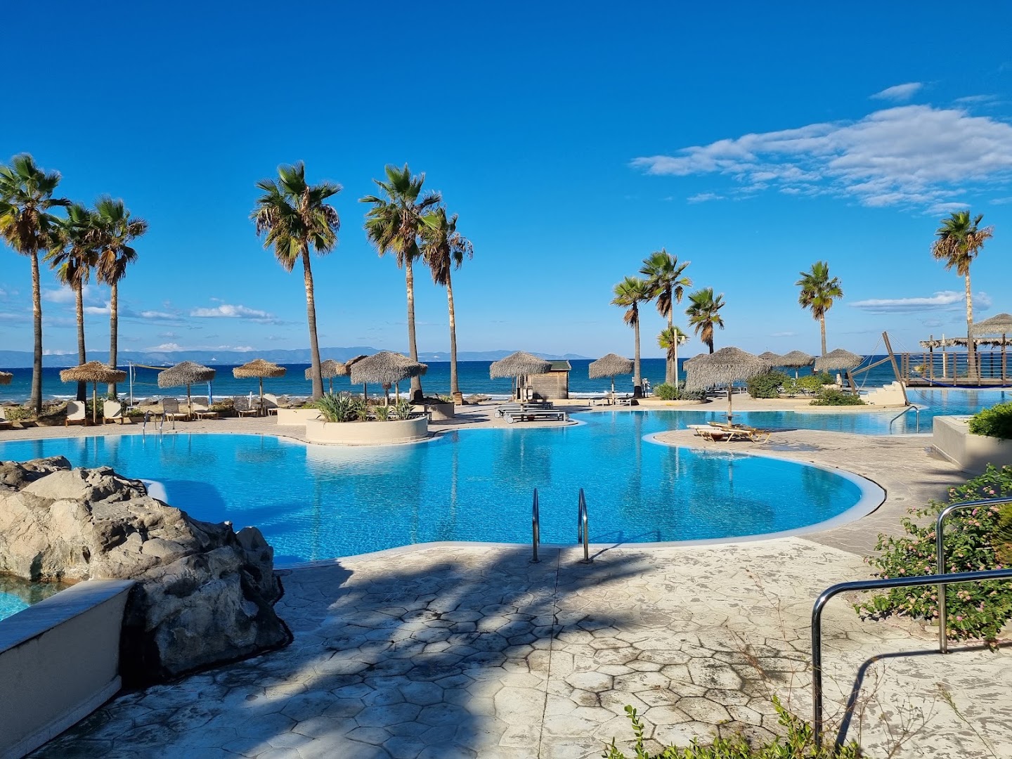 Grecotel LUXME Oasis at Riviera Olympia galvenais baseins ar skatu uz jūru un palmām — gaiša akmens terase un zilas debesis