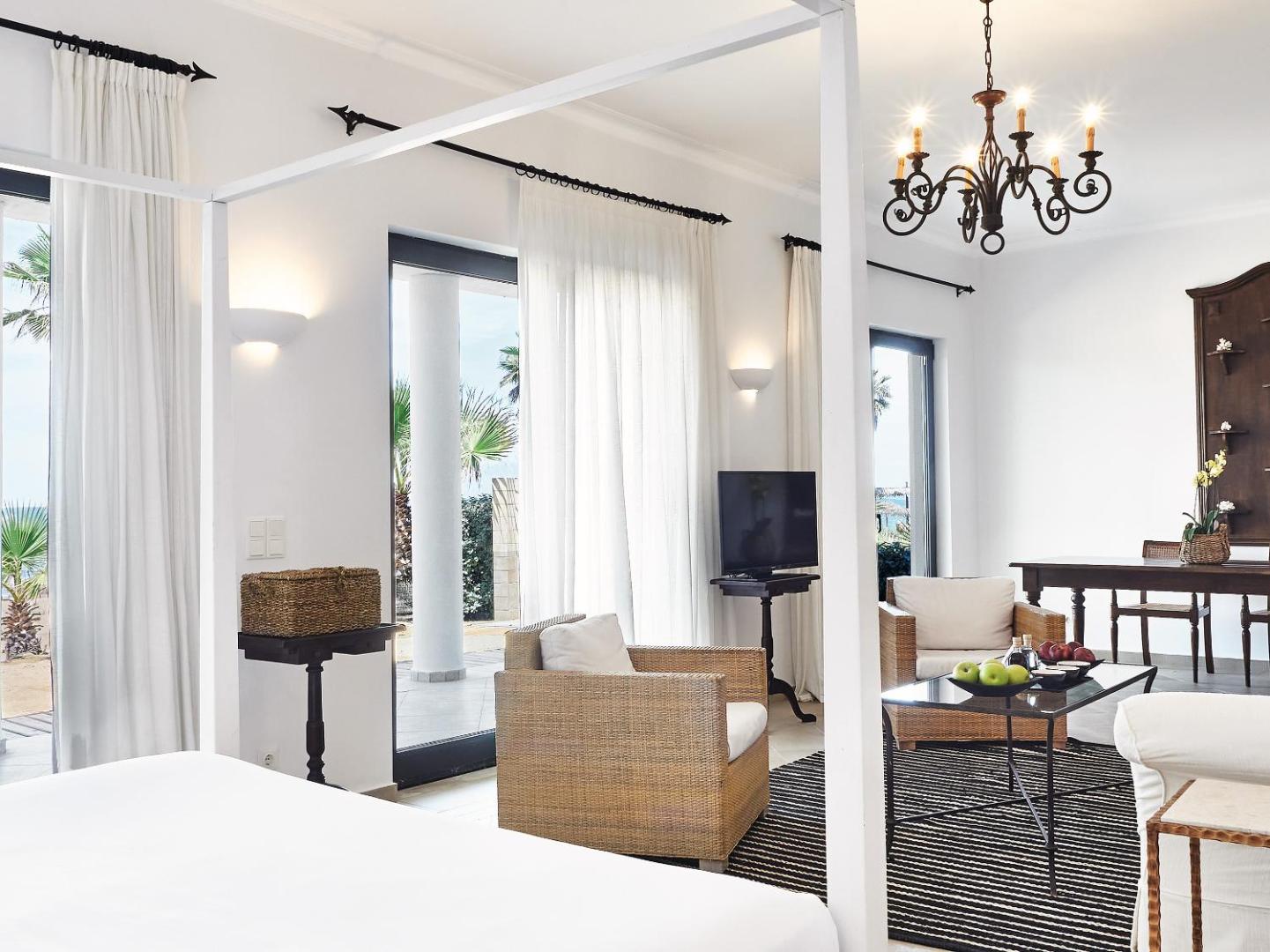 Grecotel LUXME Oasis luksusa numura dzīvojamā zona ar pītiem atpūtas krēsliem, lustru un izeju uz terasi pie jūras