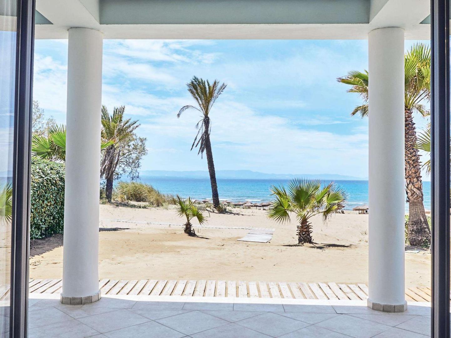 Skats no Grecotel LUXME Oasis terases uz smilšu pludmali un palmām Peloponēsas piekrastē zem skaidrām debesīm