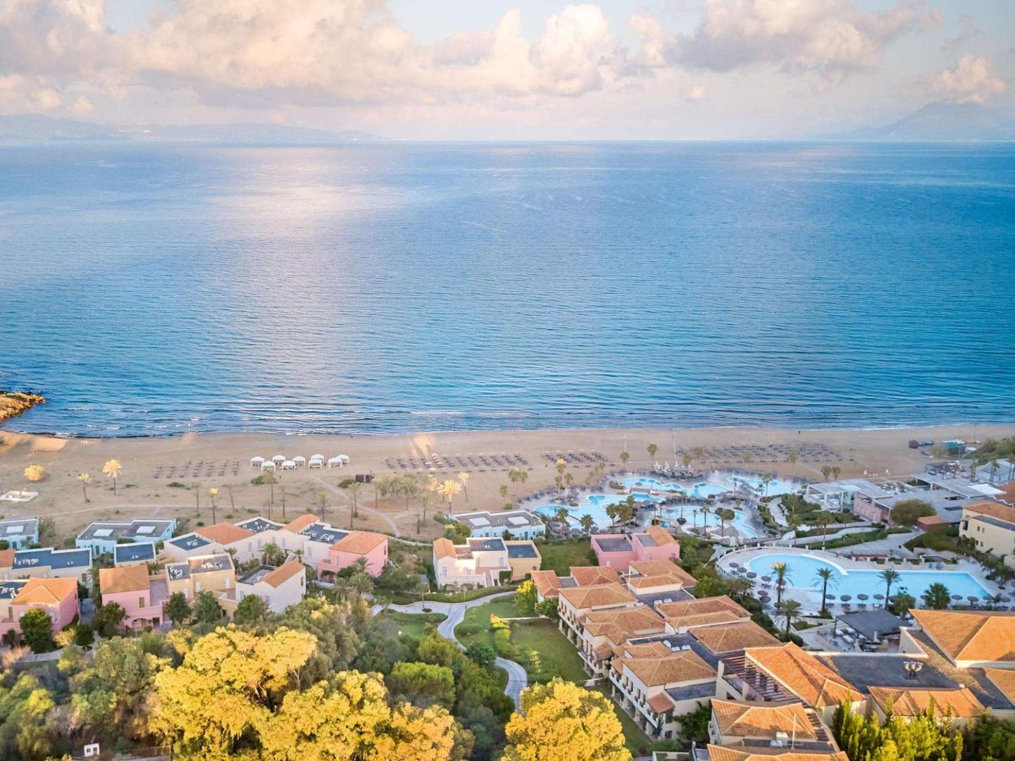 Grecotel LUXME Oasis at Riviera Olympia teritorija saulrietā ar skatu uz baseiniem, bungalo tipa numuriem un jūras horizontu.