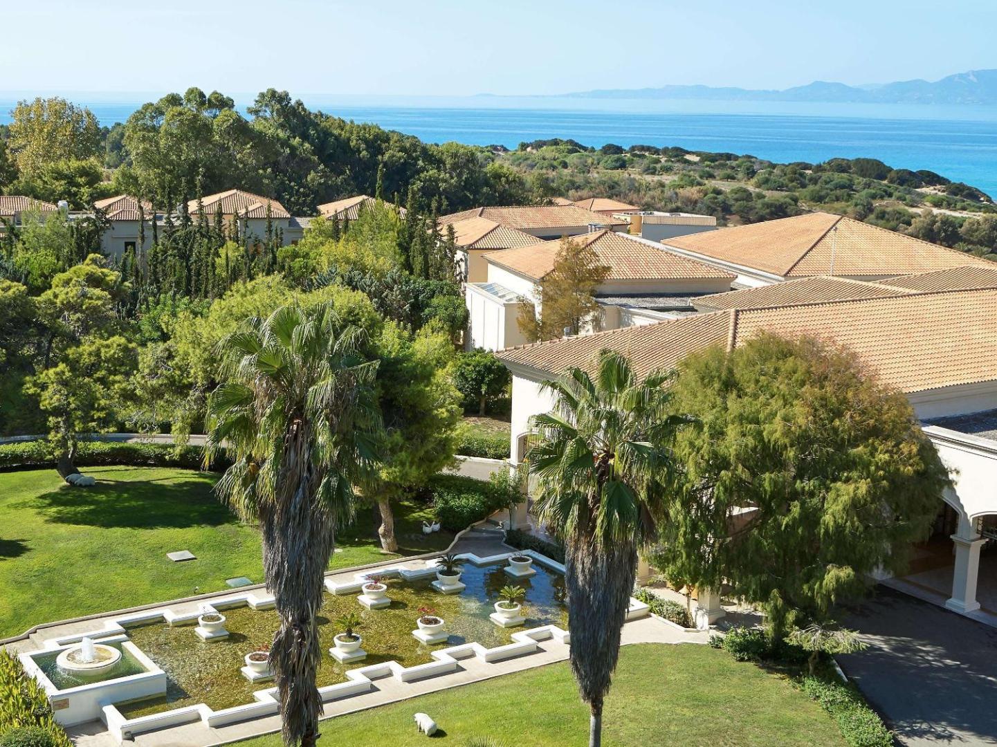 Grecotel LUXME Oasis at Riviera Olympia galvenās ēkas arhitektūra ar dekoratīviem baseiniem, palmām un skatu uz jūru fonā.