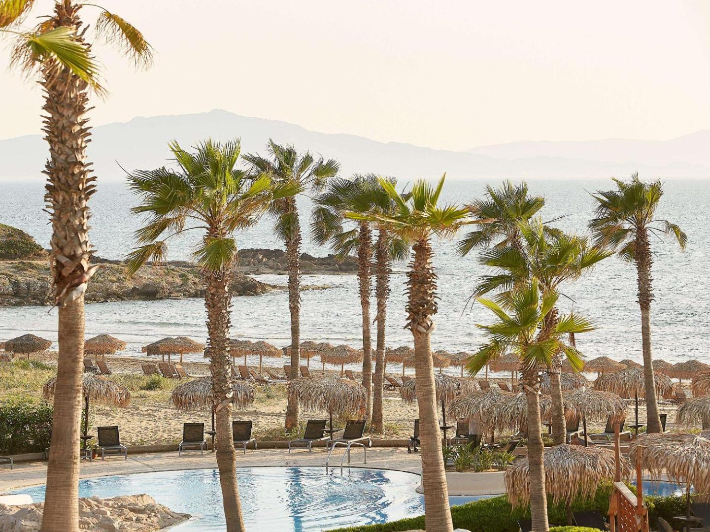 Grecotel LUXME Oasis at Riviera Olympia pludmales zona ar salmu saulessargiem un palmām — skats uz Jonijas jūru un kalniem
