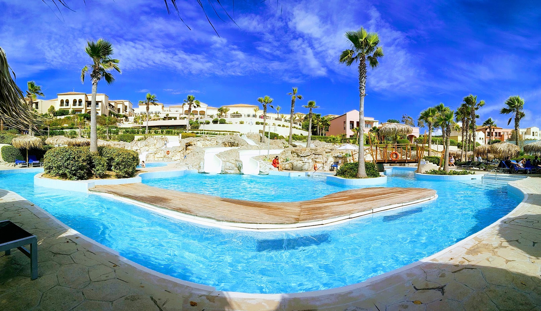 Plašs āra baseins ar koka tiltiņu un slidkalniņiem Grecotel LUXME Oasis at Riviera Olympia kūrortā Peloponēsā uz gaišu ēku fona