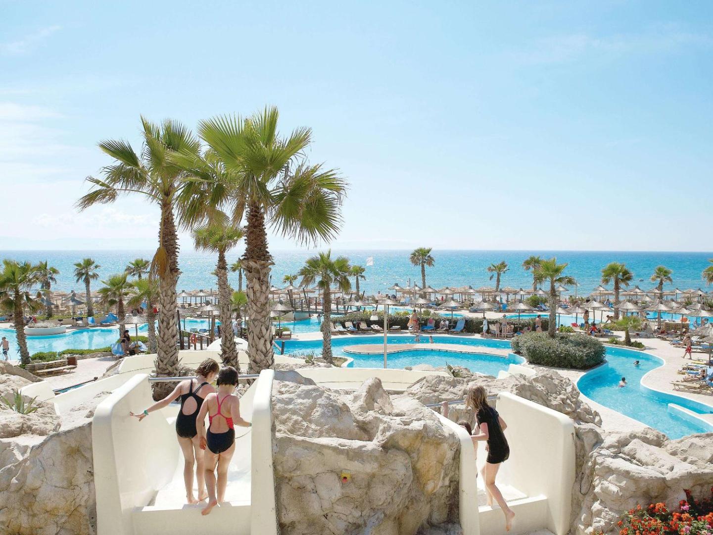Bērni uz ūdens slidkalniņiem Grecotel LUXME Oasis at Riviera Olympia akvaparkā Grieķijā — fona redzami baseini, palmas un jūra
