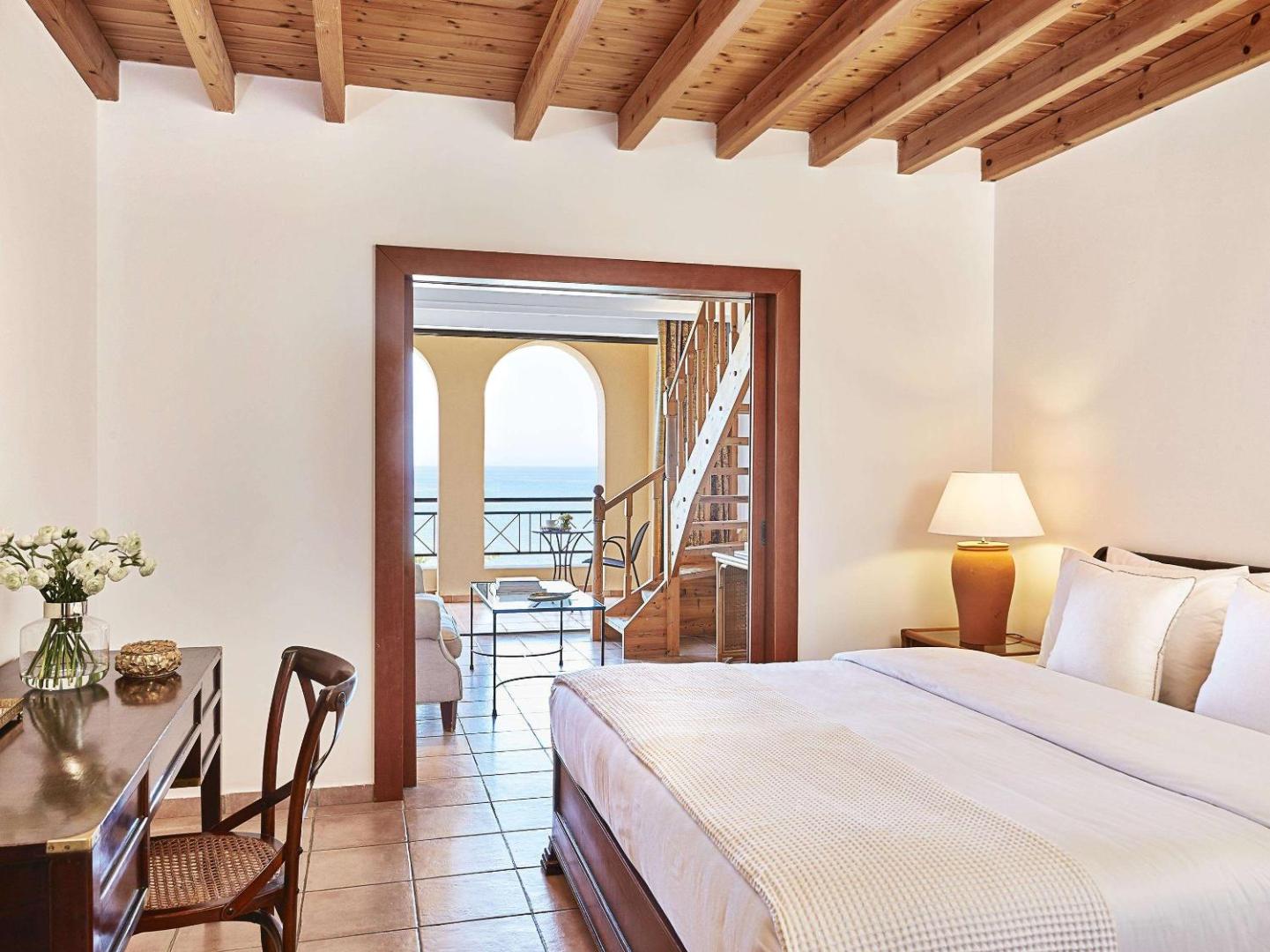 Grecotel LUXME Oasis divstāvu numura interjers ar koka siju griestiem, darba galdu un skatu uz jūru caur arkas logiem