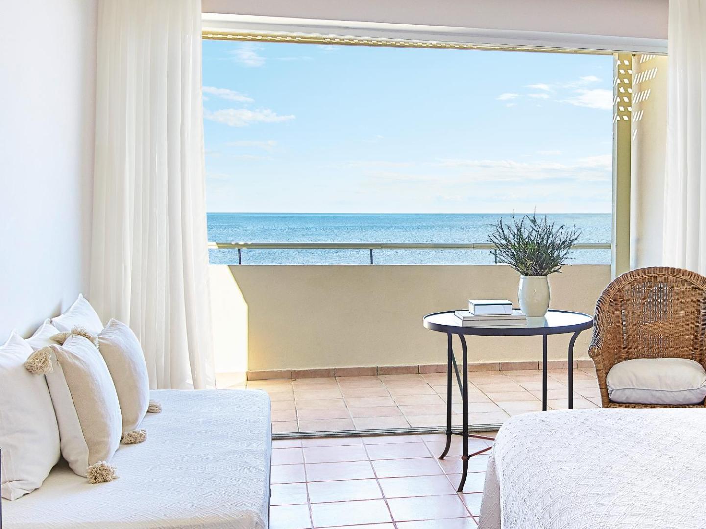 Grecotel LUXME Oasis numura balkons ar skatu uz Jonijas jūru Peloponēsā — gaišs interjers ar pīto krēslu un galdiņu