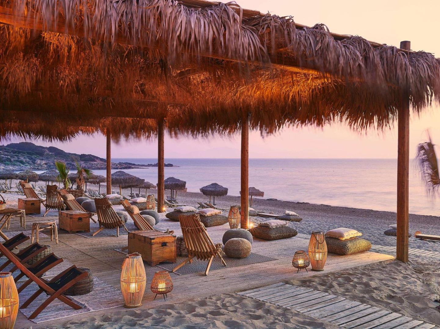 Grecotel LUXME Oasis at Riviera Olympia pludmales atpūtas zona saulrietā ar koka krēsliem, pītiem lukturiem un salmu nojumi pie jūras.
