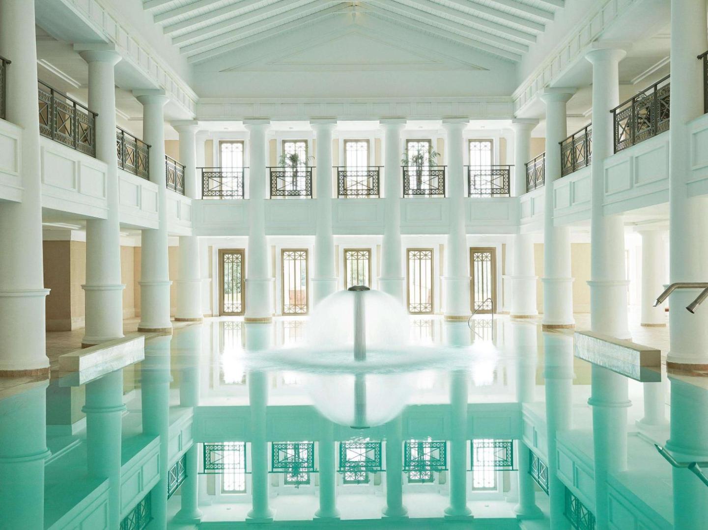 Grecotel LUXME Oasis at Riviera Olympia iekštelpu baseins ar kolonnām, strūklaku un gaišu, klasisku arhitektūru spa centrā.