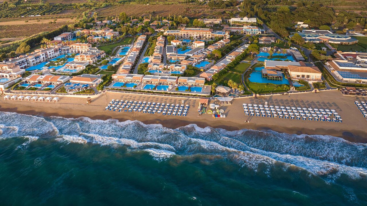 Aldemar Olympian Village kūrorta aerofoto skats no putna lidojuma Peloponēsā — plaša teritorija ar baseiniem un smilšu pludmali saulrietā