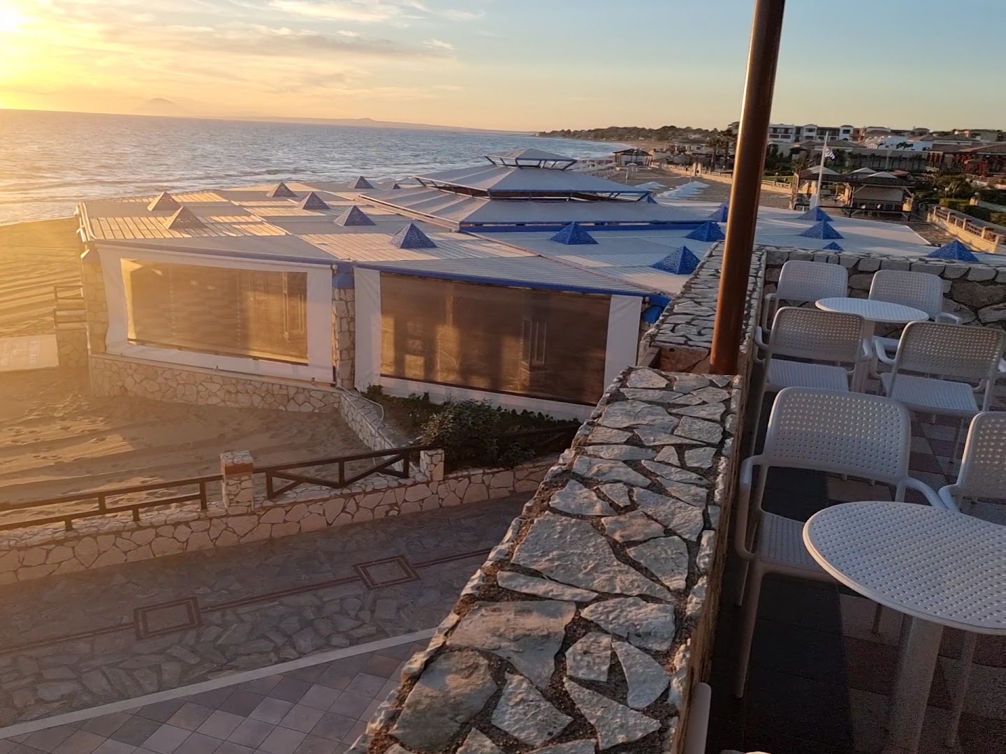 Aldemar Olympian Village pludmales bāra terase ar galdiņiem un skatu uz jūru saulrieta gaismā Grieķijas kūrortā