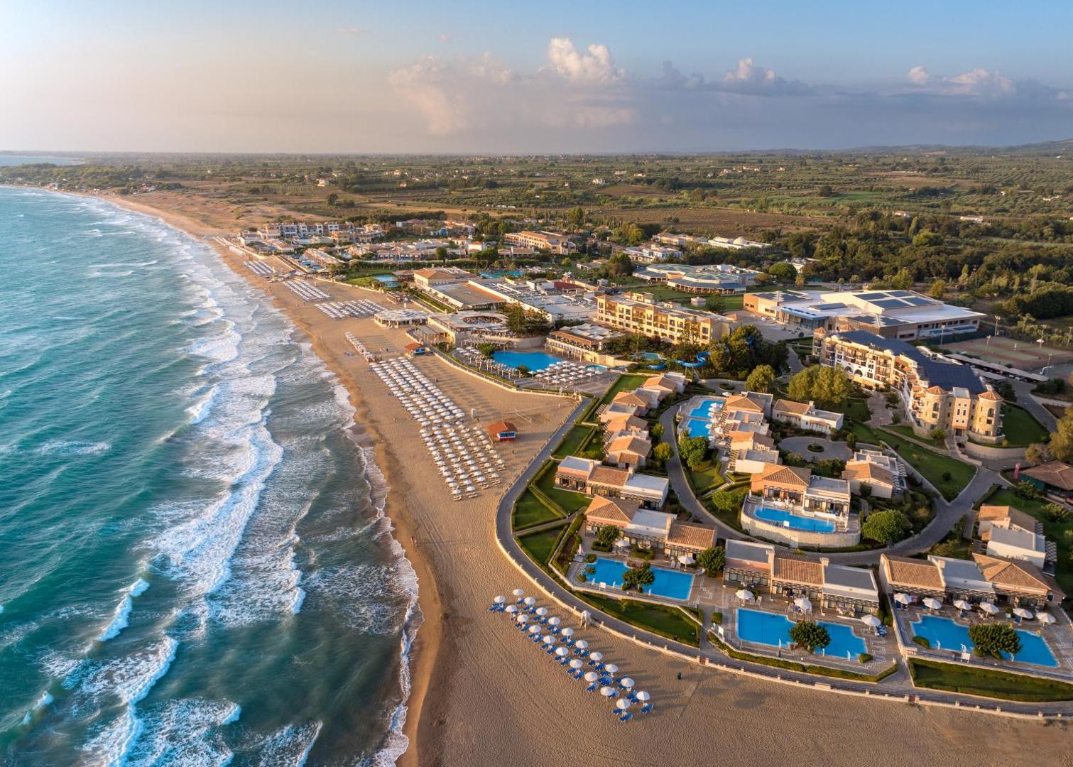 Aldemar Olympian Village viesnīcas komplekss no augšas — gara smilšu pludmale ar saulessargiem un daudziem peldbaseiniem pie numuriem