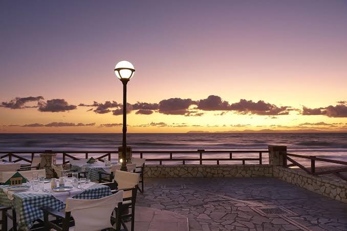 Aldemar Olympian Village restorāna terase ar galdiņiem pie jūras saulrieta laikā, skats uz horizontu un vakara apgaismojumu