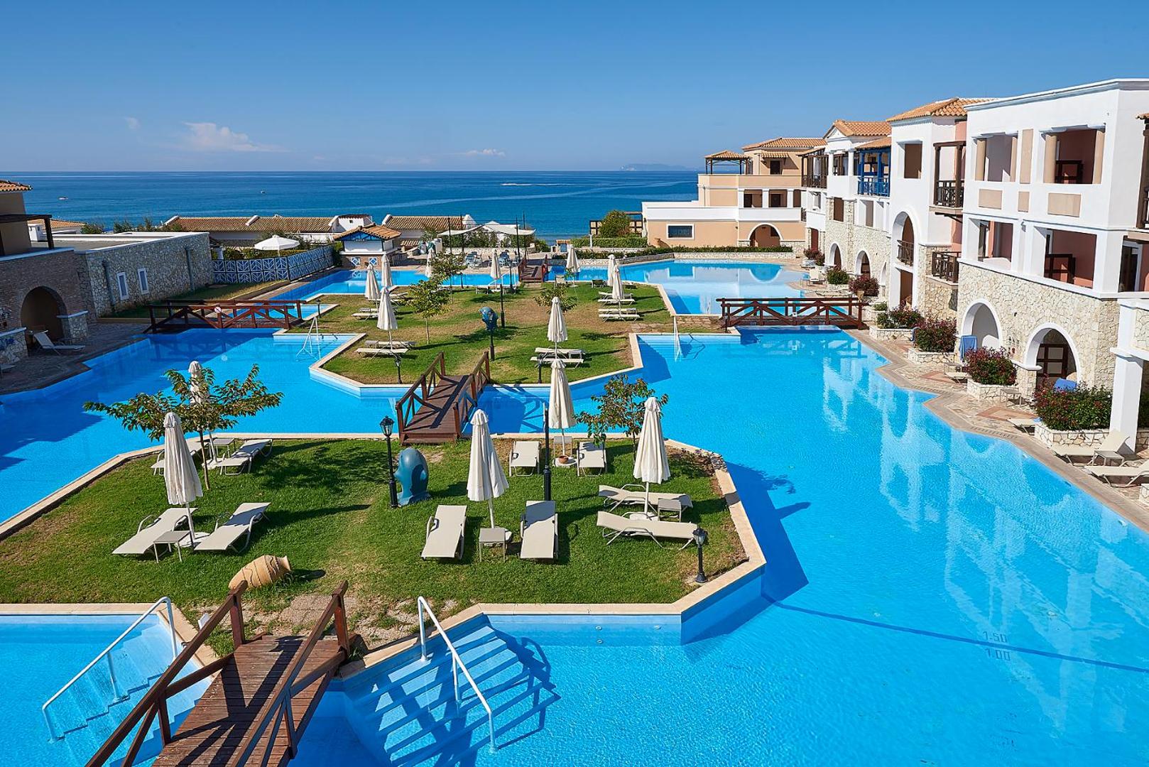 Aldemar Olympian Village kūrorta plašais āra baseinu komplekss ar tiltiņiem un sauļošanās zonām zālienā pie Jonijas jūras krasta Peloponēsā.