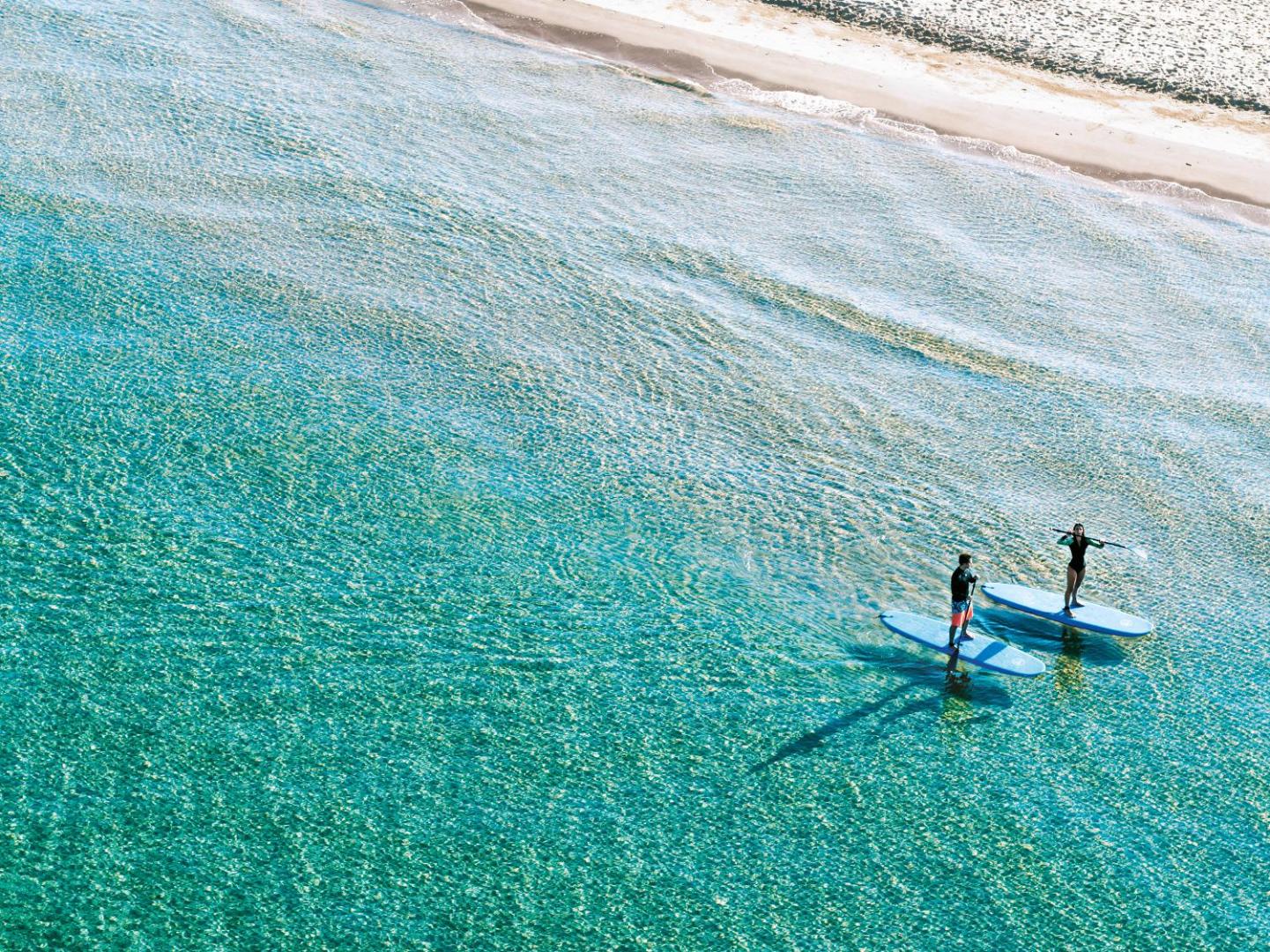 Divi cilvēki nodarbojas ar SUP sērfošanu dzidrā un tirkīzzilā Jonijas jūras ūdenī pie Grecotel Ilia Palms & Aqua Park pludmales.