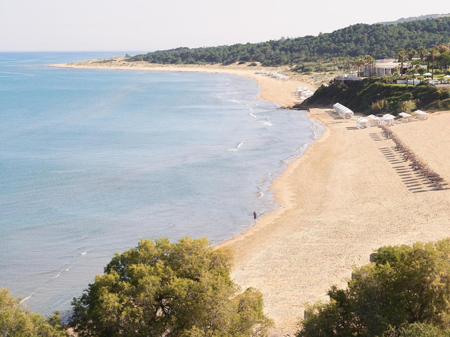 Grecotel Ilia Palms & Aqua Park plašā smilšu pludmale Peloponēsā — skats no augšas uz zilo jūru, piekrastes mežu un saulessargiem
