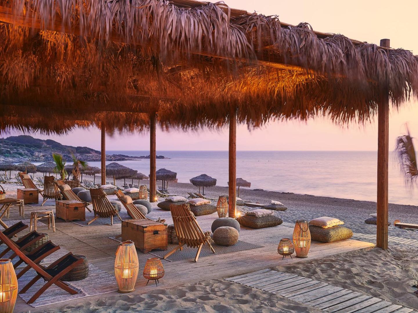 Grecotel Ilia Palms & Aqua Park pludmales bārs saulrietā Peloponēsas piekrastē — mājīga atpūtas zona ar koka krēsliem un laternām smiltīs