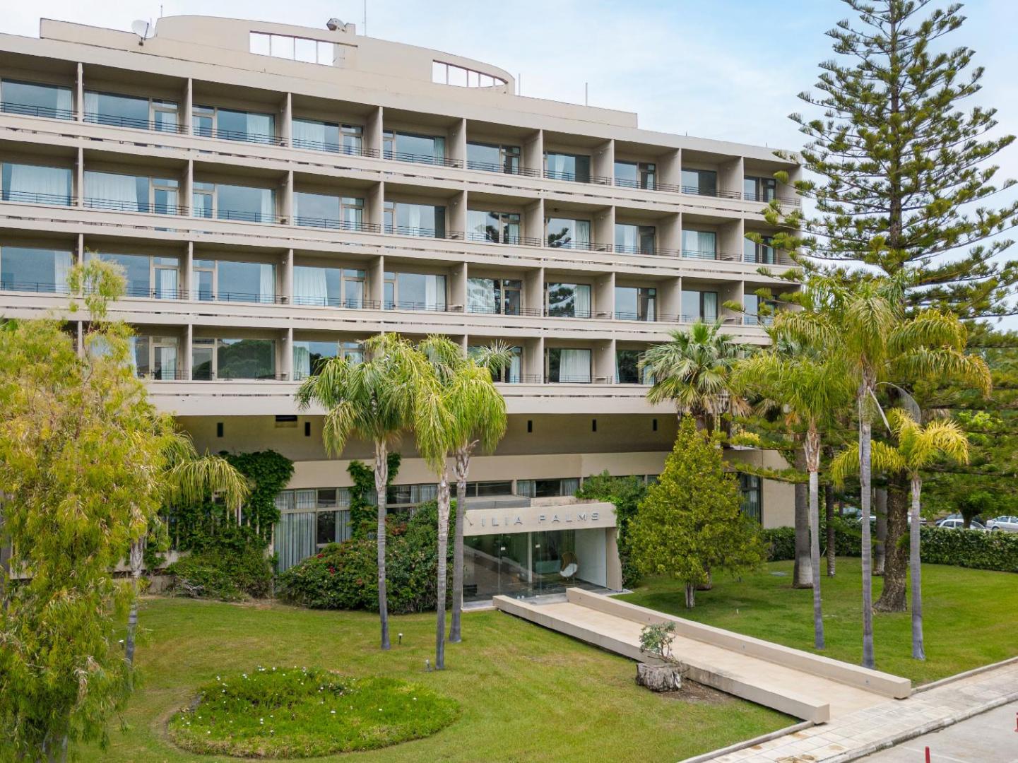 Grecotel Ilia Palms & Aqua Park galvenā ēka ar balkoniem, ko ieskauj kopti zālieni un palmas Peloponēsas kūrortā.