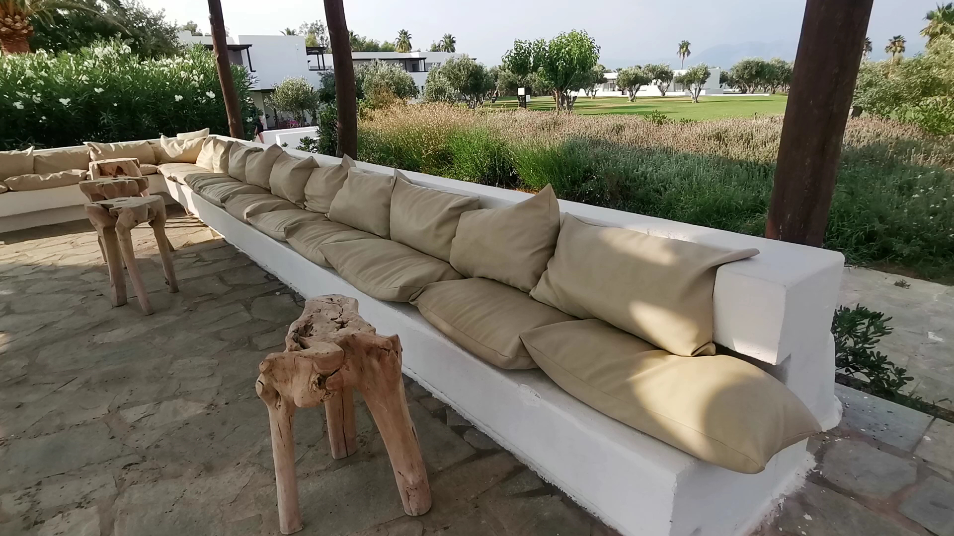 Grecotel Casa Marron atpūtas zona ar mīkstiem spilveniem un koka galdiņiem dārzā — skats uz viesnīcas teritoriju un olīvkokiem