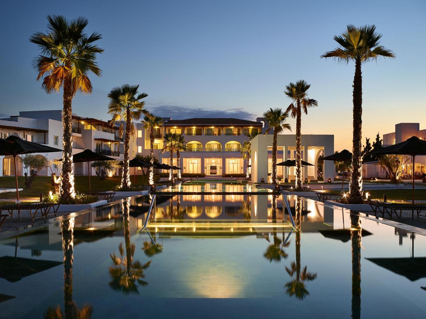 Grecotel Casa Marron kūrorta galvenā ēka un izgaismots baseins vakarā Peloponēsā, Grieķijā — palmas un atspulgi ūdenī saulrieta laikā