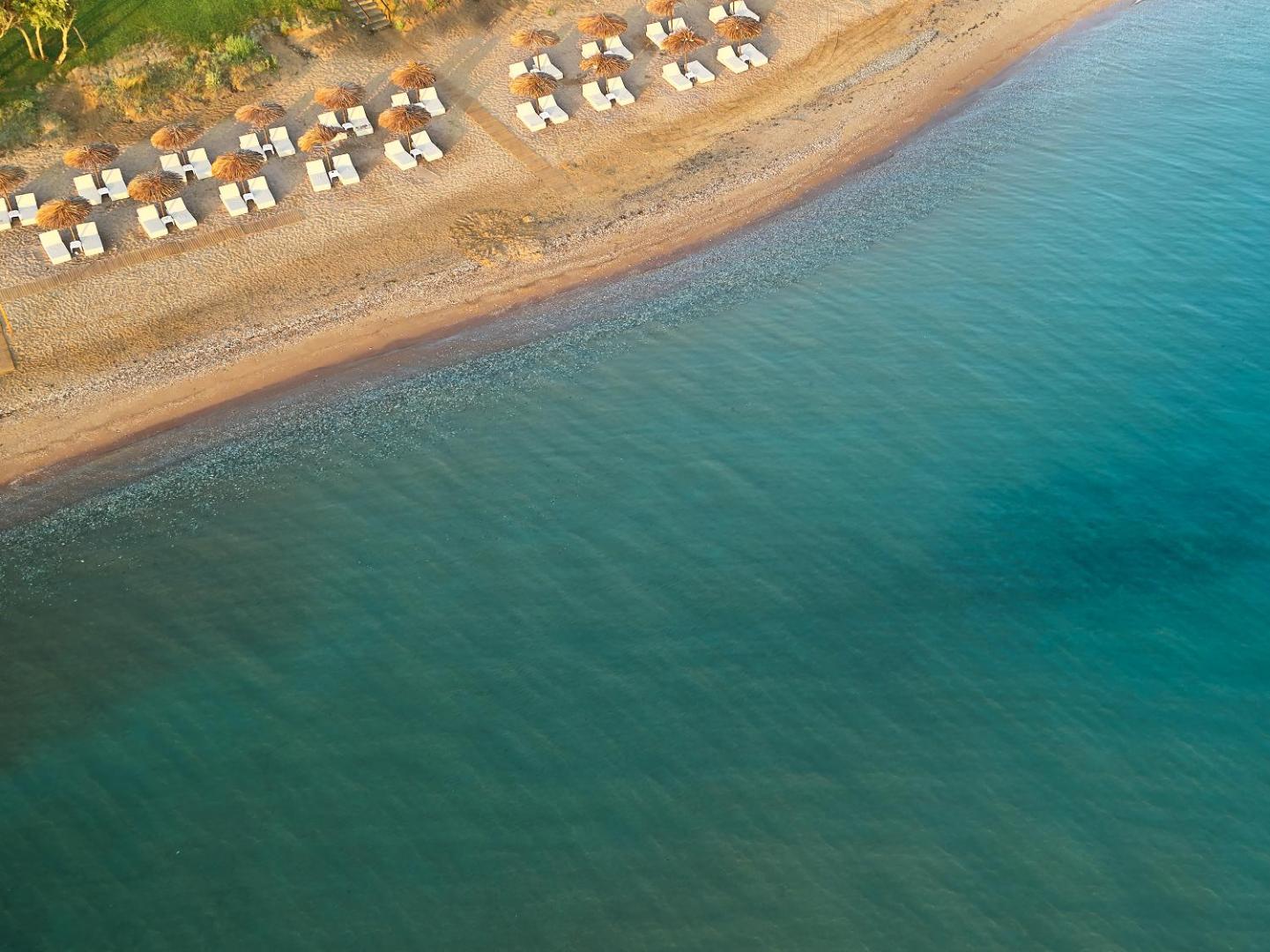 Grecotel Casa Marron aerofotoattēls ar smilšu pludmali un tirkīzzilu jūru Peloponēsā — saulessargi un sauļošanās krēsli rīta gaismā pie krasta līnijas