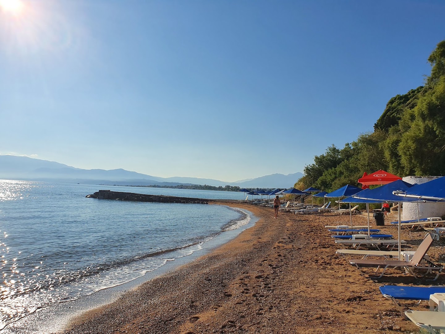 Pavlina Beach smilšu un oļu pludmale saulrietā — rindas ar sauļošanās krēsliem un skats uz kalnu grēdu horizontā