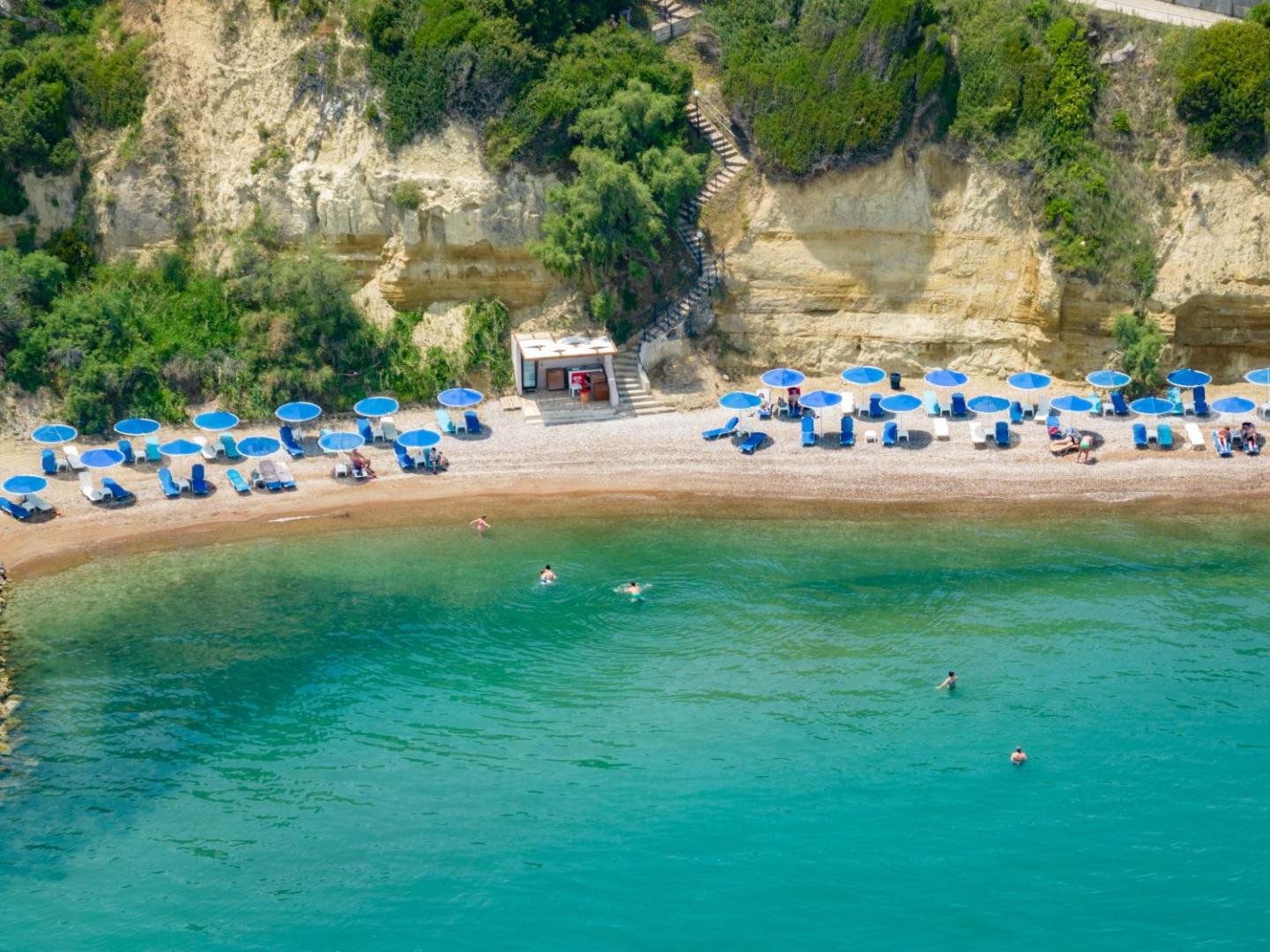 Tuvplāns uz Pavlina Beach privāto pludmali zem klints — zili saulessargi, balti sauļošanās krēsli un tirkīza krāsas jūra