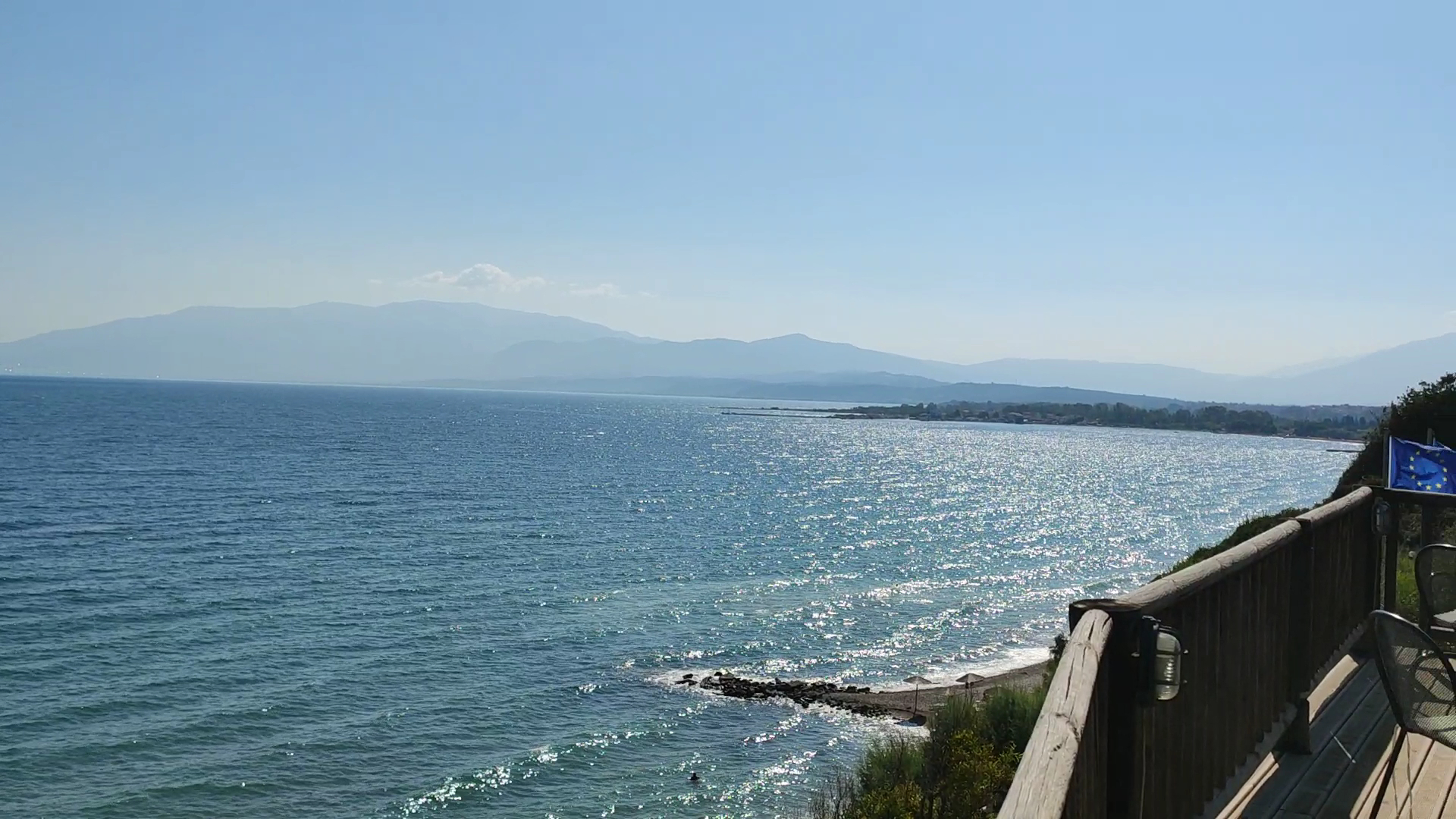 Skats no Pavlina Beach viesnīcas terases uz Jonijas jūru un kalniem tālumā — koka margas un saules pielieta piekraste