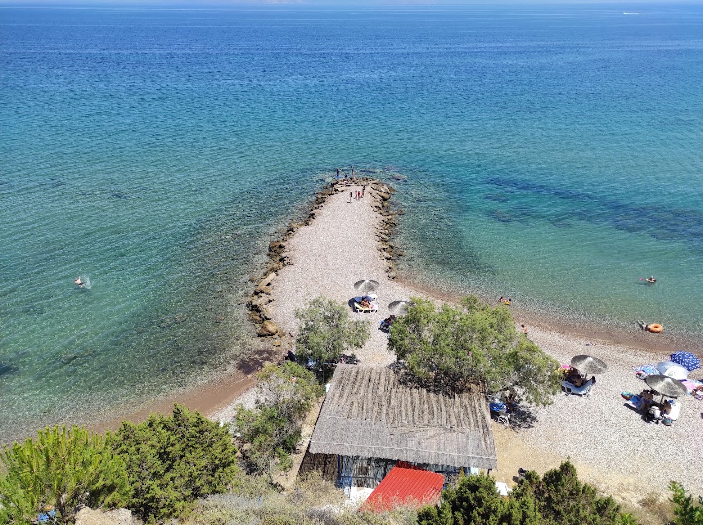 Aerofoto skats uz Pavlina Beach akmeņaino molu un kristāldzidro ūdeni — atpūtnieki pludmalē un neliela piekrastes būve