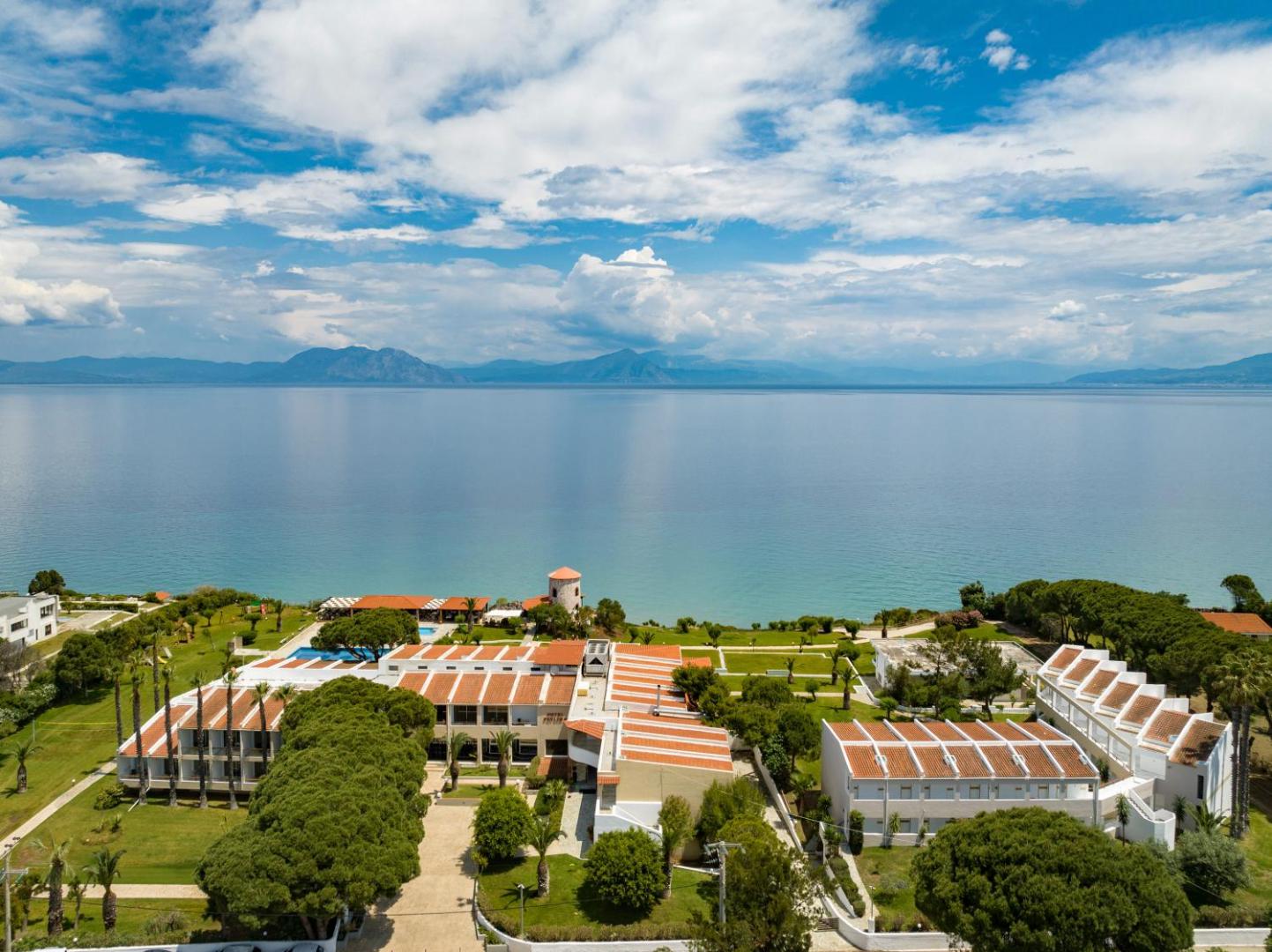 Pavlina Beach viesnīcas panorāmas skats no putna lidojuma Peloponēsas piekrastē — zilas debesis, jūra un kūrorta ēkas zaļā teritorijā.