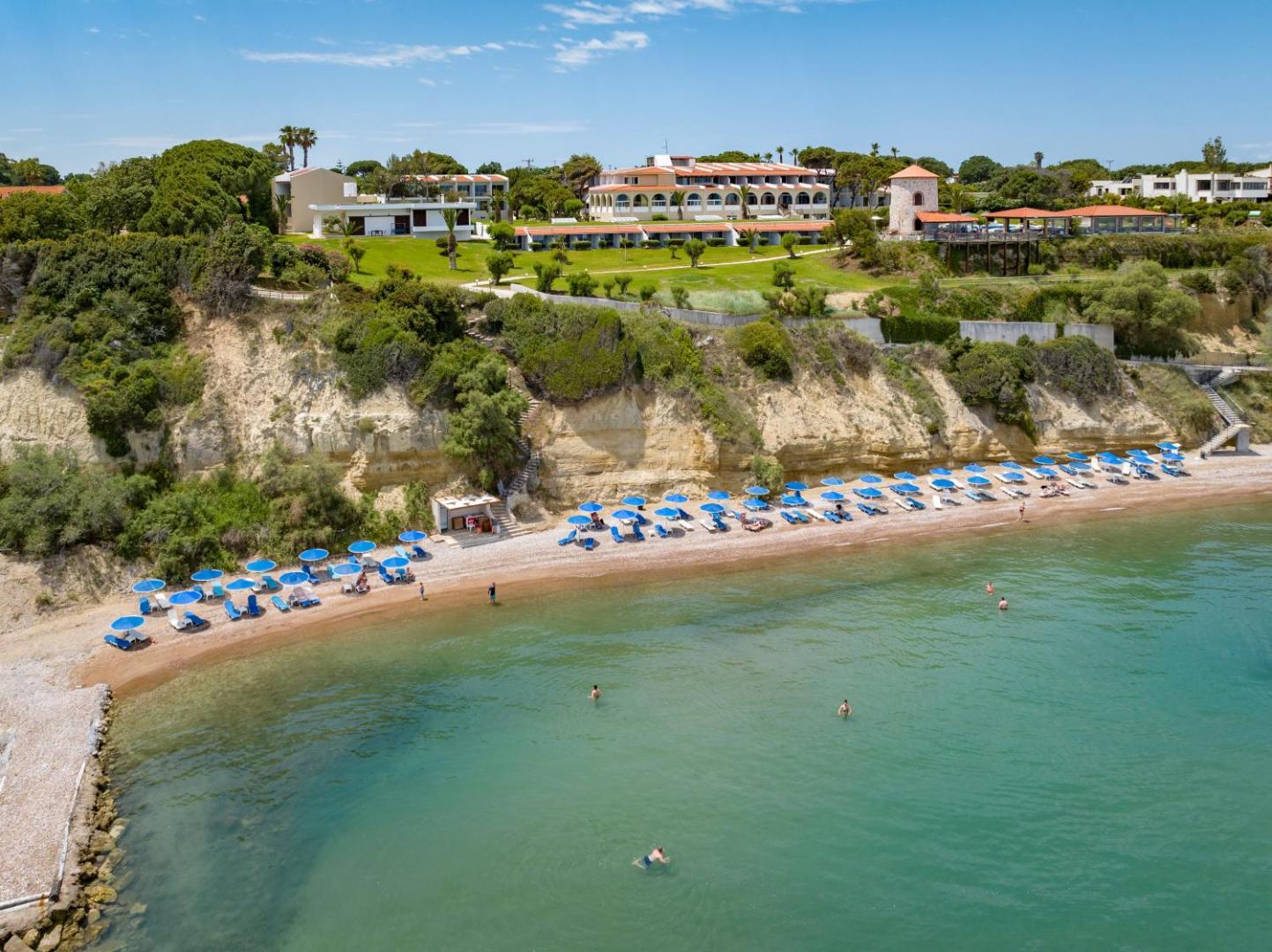 Pavlina Beach viesnīcas ēkas uz klints virs privātās pludmales Peloponēsā — zaļa teritorija, tirkīza ūdens un saulessargi