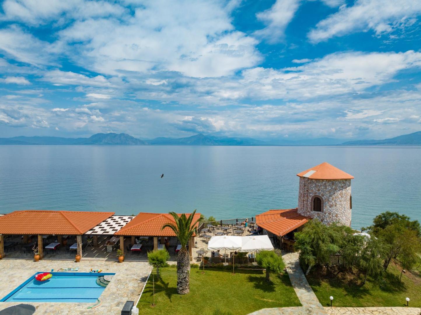 Pavlina Beach viesnīcas skats uz Jonijas jūru no piekrastes — restorāna terase, dekoratīvs akmens tornis un baseina mala Peloponēsas piekrastē