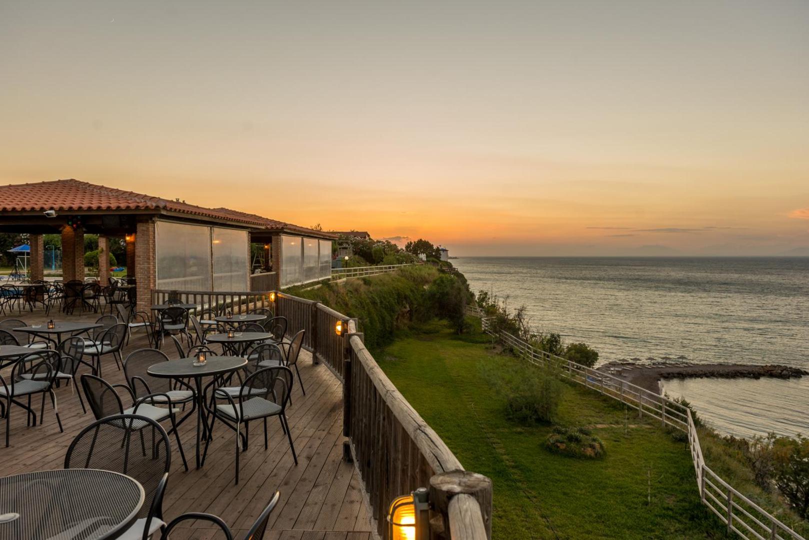 Pavlina Beach viesnīcas āra terase saulrietā ar skatu uz Jonijas jūru Peloponēsā — koka klājs, galdiņi un panorāmas ainava