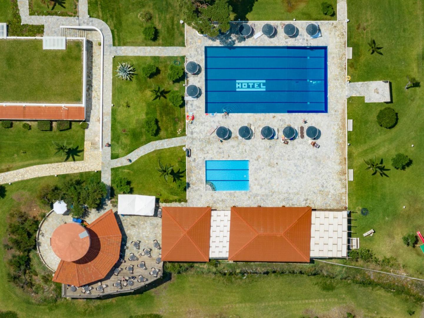 Pavlina Beach viesnīcas teritorijas skats no augšas — taisnstūra formas baseins, bērnu baseins, zaļš zāliens un terase ar sarkaniem dakstiņu jumtiem