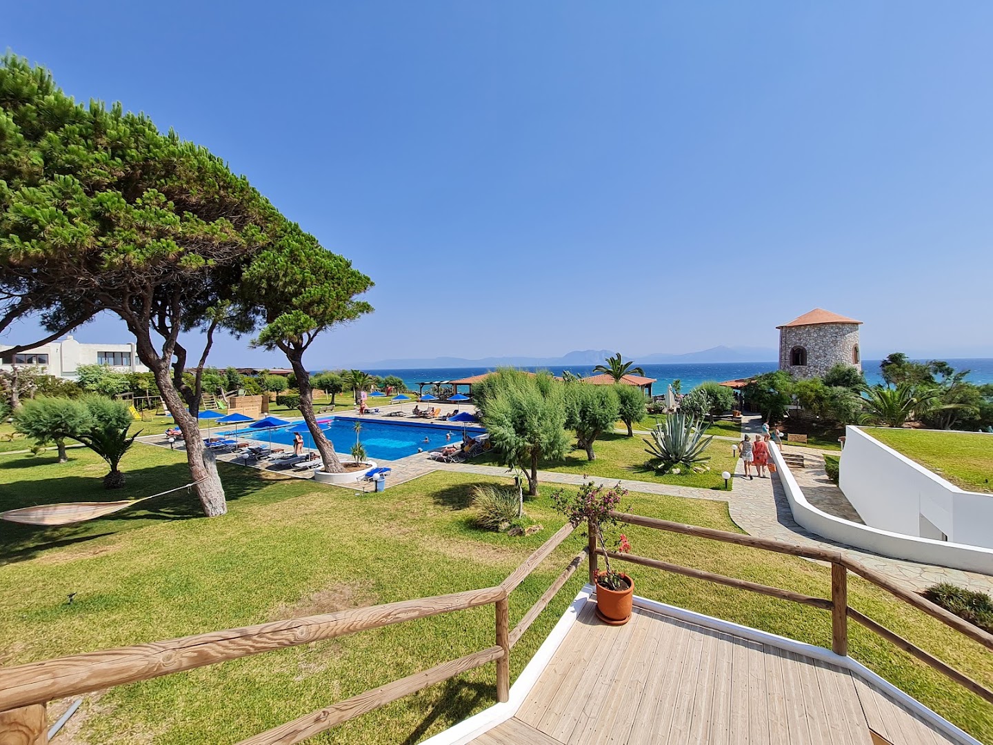 Pavlina Beach viesnīcas dārza skats no terases — zaļš zāliens ar priedēm, šūpuļtīkls, baseins un jūra tālumā Peloponēsas reģionā