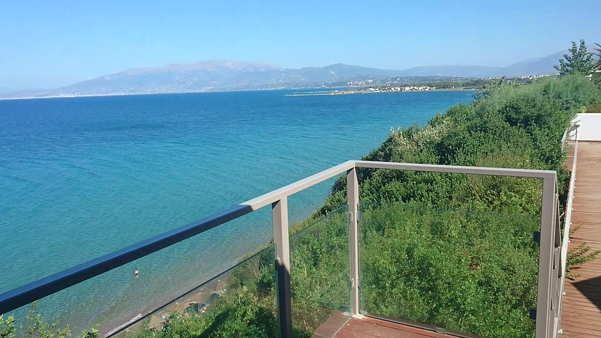 Niforeika Beach Hotel skats no terases uz Jonijas jūru un kalniem — koka klājs ar stikla margām un piekrastes zaļumiem