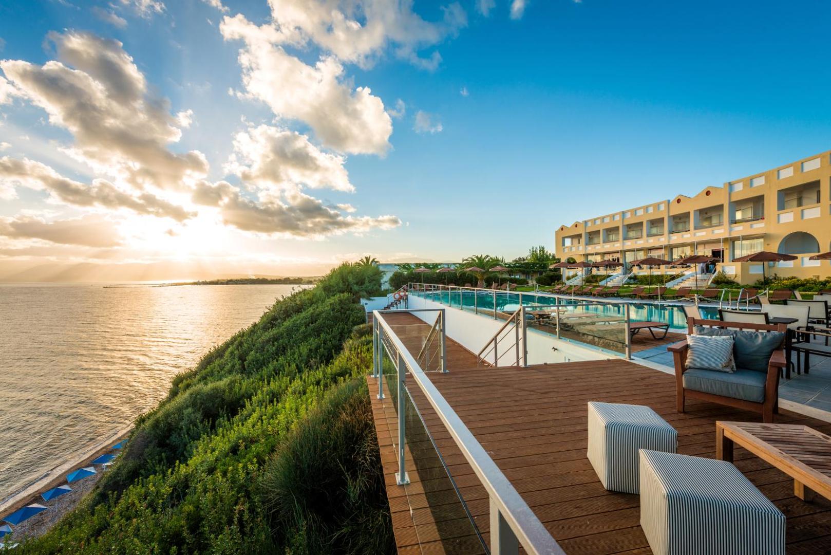 Niforeika Beach Hotel & Bungalows terase ar skatu uz jūru saulrietā Peloponēsā — koka klājs, atpūtas mēbeles un baseins piekrastē